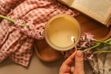 Hand lighting a candle next to a cup of tea, gift box, and pink hyacinths on a cozy fabric, creating a warm and peaceful springtime flat lay