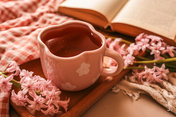 Pink flower-shaped mug filled with tea on a wooden tray with pink hyacinths, gingham fabric, and an open book, creating a cozy springtime reading scene