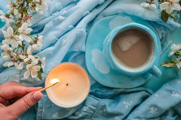 A hand lighting a candle with a match, with white spring blossoms and a blue cup of coffee in the background, creating a warm, peaceful, and cozy scene