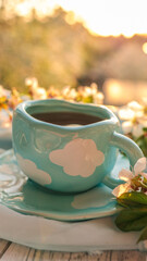 Pastel blue ceramic mug with white cloud pattern on matching saucer, surrounded by cherry blossoms in soft sunset light, creating a cozy spring atmosphere
