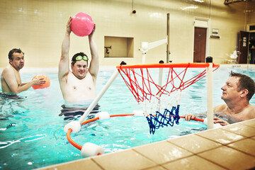 Disability, fitness and swimming activity by down syndrome man playing basketball in indoor pool. Care facility for special needs people training in water, having fun and bonding in physical therapy