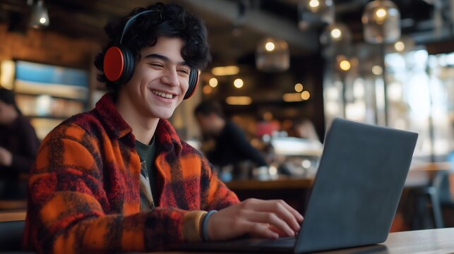 Young man with curly dark hair wearing red headphones and plaid flannel shirt smiling while using a dark laptop in a cozy coffee shop with warm lighting and blurred background atmosphere