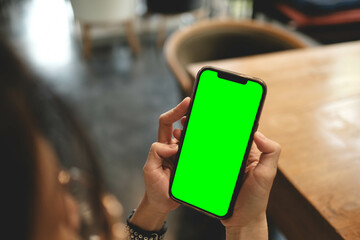 Close-up of woman holding a smartphone with green screen while sitting in a cozy cafe setting.