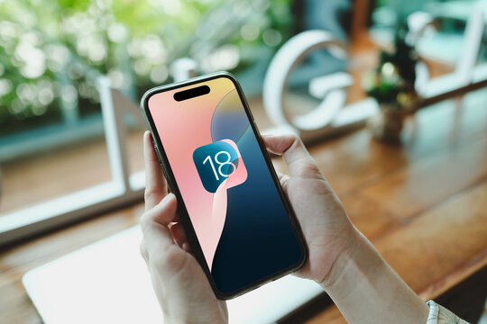 Bangkok, Thailand. May 1, 2025; User holding smartphone displaying colorful iOS 18 update screen with blurred greenery background.