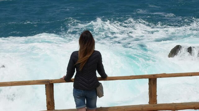 A woman with her back turned and long dark hair admires the waves crashing against the coast of Fuerteventura. In the Ajui caves.