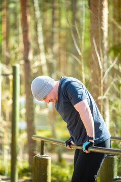 Caucasian man training outdoors on parallel bars in sunny forest park. Wearing a beanie and gloves he performs bodyweight dips, showcasing strength, fitness and healthy lifestyle in public park
