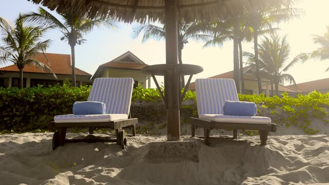 Relaxing tropical beach chairs under straw umbrella with sunlit resort background
