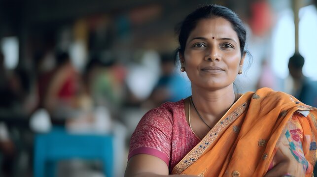 Indian woman in an embroidered pink blouse and vibrant orange floral sari, wearing a traditional bindi and looking serenely at the camera in a colorful local market.