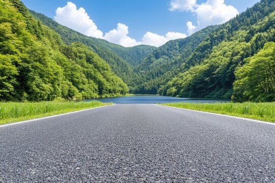 Empty asphalt road leading to a serene lake surrounded by lush green forests under a partly cloudy sky