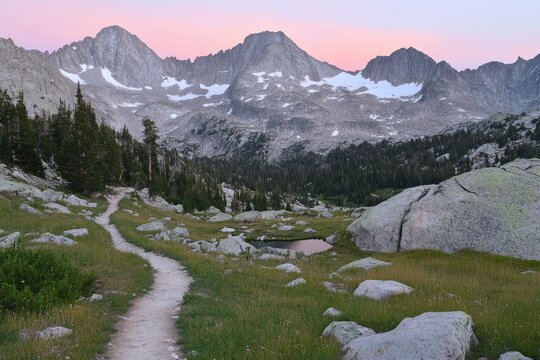 Mountain path winds through a meadow, towards a serene alpine lake nestled beneath a range of granite peaks bathed in the soft light of dawn or dusk