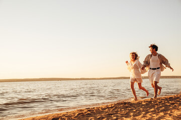 Smiling beautiful woman and her handsome boyfriend. Excited couple in casual clothes. Happy cheerful family. Female and man run at sunrise over sea beach outdoors. Run at seaside in summer day