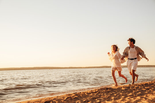 Smiling beautiful woman and her handsome boyfriend. Excited couple in casual clothes. Happy cheerful family. Female and man run at sunrise over sea beach outdoors. Run at seaside in summer day - Powered by Adobe