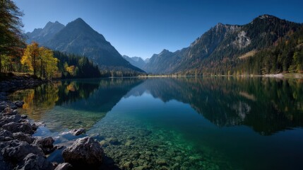 Fototapeta premium Crystal Clear Lake Reflects Towering Mountains Under A Bright Blue Sky Featuring Rocky Shores Dense Forests And Deep Green Water In A Scenic Landscape