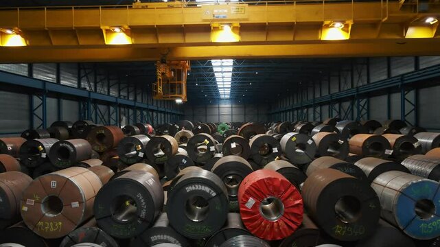 A large overhead bridge crane moves towards the camera in a warehouse filled with steel coils