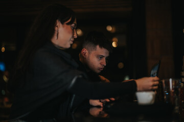 Two individuals collaborate on a laptop in a dimly lit cafe environment, showcasing teamwork and...