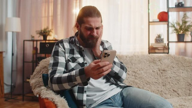 Man at home pressing cheek in hard tooth pain, quickly dialing phone for immediate dentist contact. Guy on sofa shows frustration and tension, suffer from painful toothache, sensitive teeth, cavities