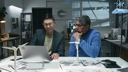Medium shot of young and mature diverse engineers sitting at table with laptop and working on new digital wind turbine model in office, multiple technical drawings and two windmill maquettes on desk