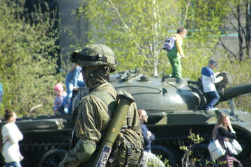 Military soldier observes visitors near tank during outdoor event in a public park setting on a sunny day