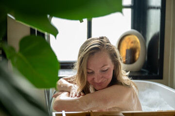 Woman relaxing in bathtub near window with plants and candles around her