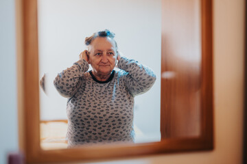 An elderly woman stands in front of a mirror, adjusting her hair, wearing a patterned shirt. The...