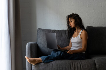 Young woman sitting barefoot on a sofa, working on a laptop with natural window light in a...