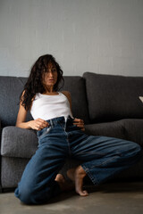 Fototapeta premium Confident young woman with tousled hair sitting on the floor in front of a grey couch, wearing a white tank top and blue jeans, looking directly at the camera in soft indoor light.