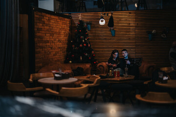 Two individuals relax in a warmly lit cafe, sitting on a sofa near a beautifully decorated Christmas tree, creating an intimate and festive setting for sharing moments or working together.