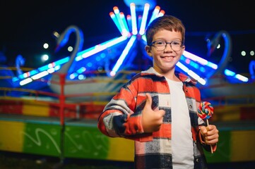 A cute caucasian boy eating a colorful lollipop standing against the background of a carousel in...