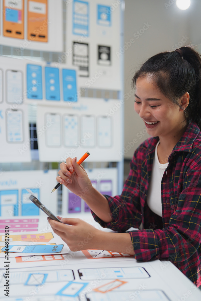 Wall mural smiling female web designer engaging with her cellphone and pen while creating a mobile application 