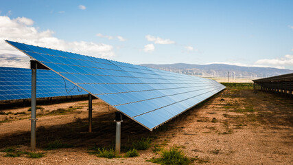Close-up angle of modern solar panels in renewable energy facility with clear sky and wind turbines across dry, semi-arid terrain near mountains