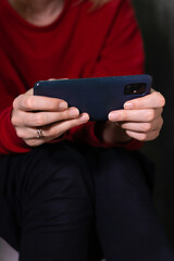 Woman wearing a vibrant red sweater and dark pants holds a smartphone horizontally in both hands, sitting on the toilet. Vertical photo