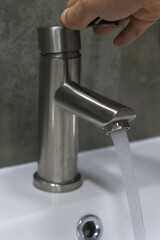 Hand turning on a brushed nickel faucet in a modern bathroom, with clear water flowing into a clean white sink, promoting water conservation and hygiene. Vertical photo