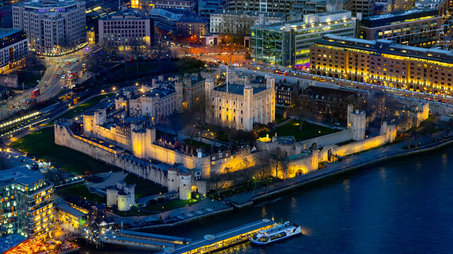 A vibrant aerial view captures the illuminated Tower of London at dusk. The historic castle stands majestically beside the Thames, surrounded by the sparkling city lights of London.