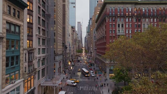 Cars and buses drive along a busy Fifth Avenue near Madison Square Park, flanked by tall buildings of Flatiron District, as seen from a high vantage point at dusk