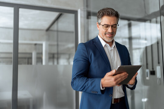 Focused senior mature Indian or Latin entrepreneur businessman holding digital pc tablet standing in office. Smiling hispanic man in suit working using touchpad computer for business work project