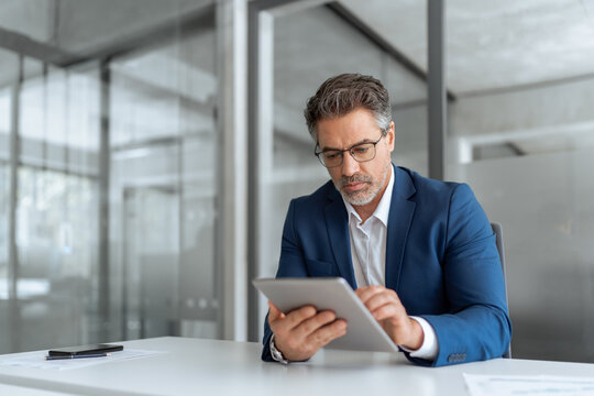 Concentrated executive senior man in suit working using computer for business banking work. Focused senior mature ceo manager businessman looking at digital tablet screen at desk in modern office