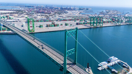 High-angle view of the iconic Vincent Thomas Bridge spanning the Port of Long Beach in California. The turquoise suspension bridge crosses over calm blue water, with stacks of shipping container