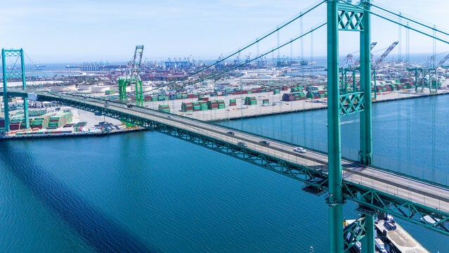 High-angle view of the iconic Vincent Thomas Bridge spanning the Port of Long Beach in California. The turquoise suspension bridge crosses over calm blue water, with stacks of shipping container