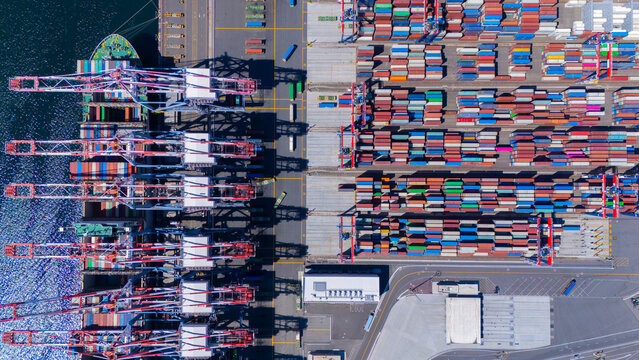 A straight-down aerial photo of shipping cranes operating over a dock filled with colorful cargo containers. The image highlights the symmetry and industrial activity of a modern shipping port.