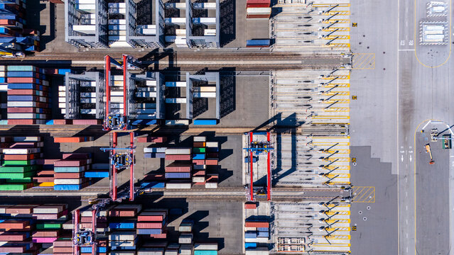 A straight-down aerial photo of shipping cranes operating over a dock filled with colorful cargo containers. The image highlights the symmetry and industrial activity of a modern shipping port.