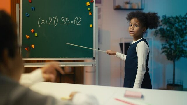 Funny girl playing strict math teacher, pointing at chalkboard during lesson