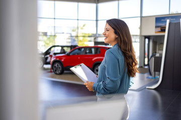 Portrait of pretty Caucasian vehicle saleswoman in suit standing with clipboard near new modern car in bright and spacious showroom. Professional female salesperson at car dealership.