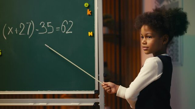 Young African American girl with pointer explaining math problem near chalkboard