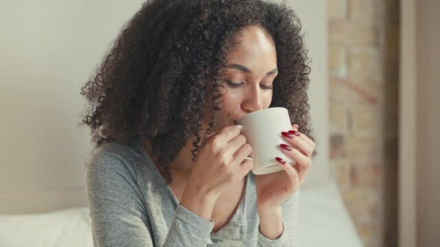 Elegant African American woman holding ceramic mug and gently taking sip of hot drink. Enjoying cozy morning while looking ahead with thoughtful expression in softly lit bedroom corner.