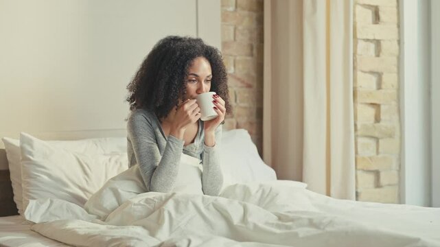 Content African American female sitting in bed and holding white mug with both hands. Tasting warm drink while watching outside window and enjoying cozy peaceful morning in modern bedroom.
