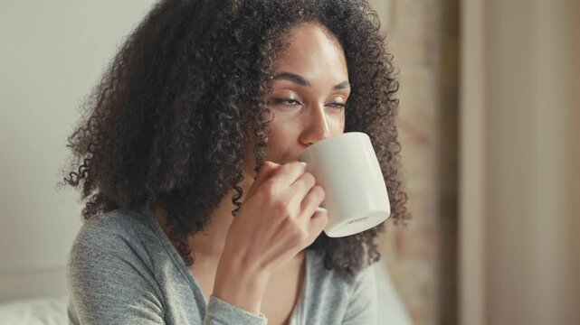 Radiant African American woman smiling warmly while holding mug in hands. Taking sip of drink. Appearing cheerful and refreshed while enjoying pleasant morning in peaceful bedroom setting.