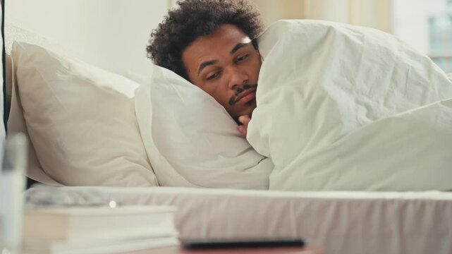 Sleepy African American male just waking up while lying under blanket in bed. Picking up smartphone with tired eyes, checking notifications early in the morning. Starting day passively.