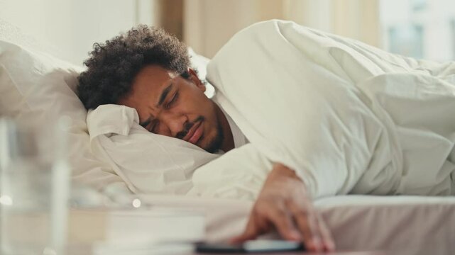 Sleepy African American male turning off phone alarm while lying in bed under blanket. Eyes barely open, reaching out lazily with groggy expression, preparing to fall back asleep after distraction.