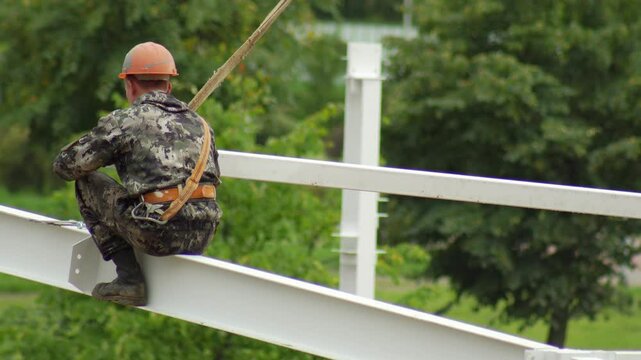 A worker fitter in a protective suit and helmet assembles a metal structure at a construction site in summer, industry. Copy space for text
