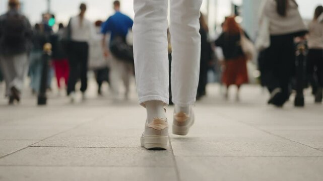 Back view following shot female feet steps walking pedestrian crossing street, sunny day, young woman wearing white pants and sneakers goes crosswalk. Low angle people legs stepping zebra asphalt